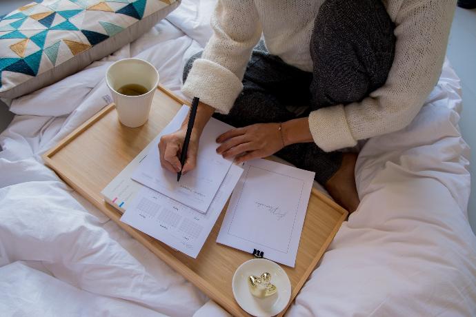 person in white sweater writing on white paper beside white ceramic mug on brown wooden table